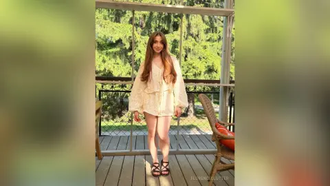 Young woman in a cream bohemian blouse standing on a sunny wooden porch surrounded by lush green trees.