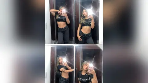 Young woman in a black crop top and dark jeans taking a mirror selfie in a modern indoor setting.