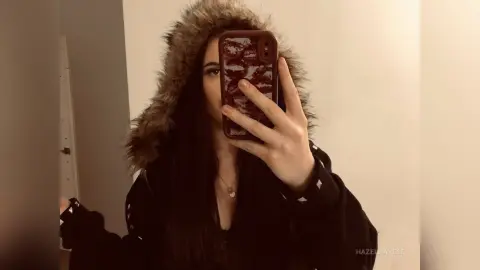 Young woman in a black hooded jacket with faux fur trim taking a moody mirror selfie with a patterned phone case.