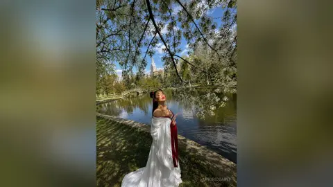 IngridSaint poses in a white dress by a lake under the shade of large trees.