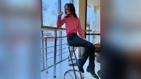 Stylish woman in a red striped shirt and dark jeans sitting on a bar stool in a bright modern interior.