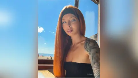 Redheaded woman in a black tube top posing outdoors against a bright blue sky background.