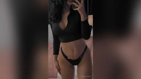 Curvy woman in black crop top and bikini bottoms taking a mirror selfie in a moody bedroom setting.