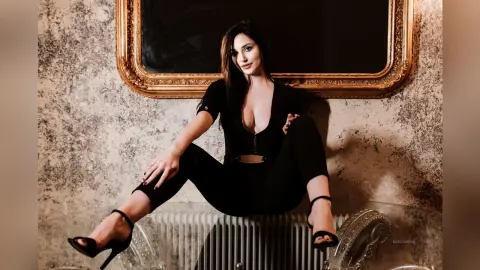 Elegant woman in a black jumpsuit sitting against a textured wall near a gold vintage mirror.