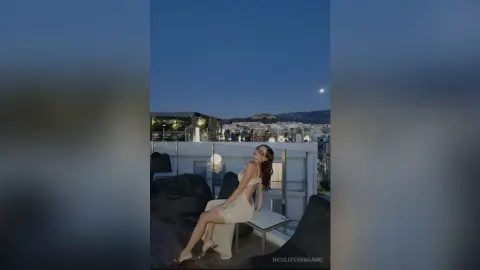 Elegant woman in a white evening gown sitting on a rooftop terrace overlooking a city skyline at twilight.