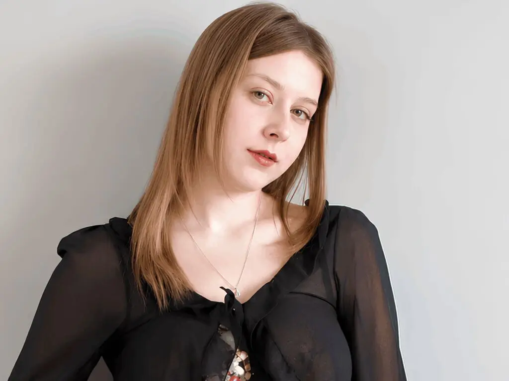Portrait of a young woman with long blonde hair wearing a black blouse against a neutral grey background.