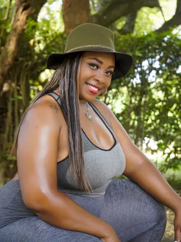 Smiling Black woman in a grey tank top and olive fedora relaxing in a lush green garden setting.