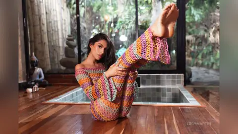 LucyMartin sits on a wooden floor by a small pool, wearing a colorful, patterned off-the-shoulder long-sleeve top and matching bottoms while lifting one bare leg high.