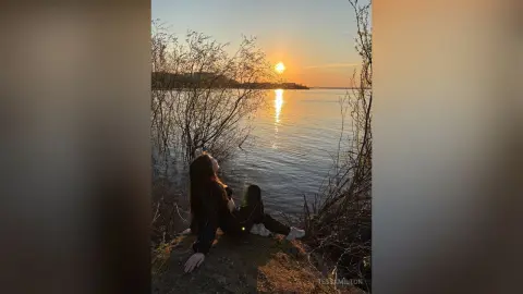 TessaMilton sits on a rocky shore by the water, looking up at the sunset through the trees.