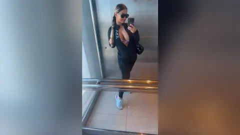 Smiling woman in black athletic leggings and jacket taking a mirror selfie in a modern elevator.