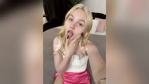 LillyEyes licks her lips while wearing a white tank top and pink satin skirt, posing on a bed with her blonde hair down.