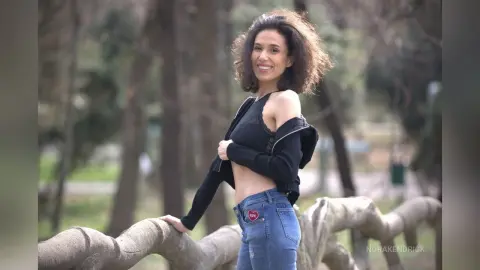 Smiling woman in a black crop top and blue jeans posing outdoors in a park setting.