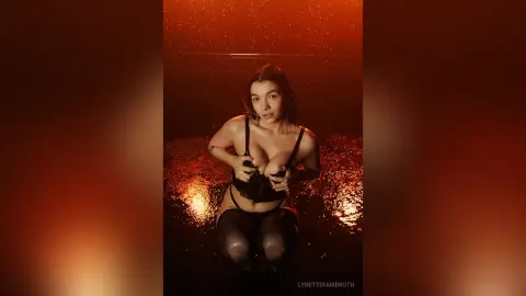 Young woman in black lace lingerie and stockings posing in a moody, warm-lit setting with golden bokeh lights.