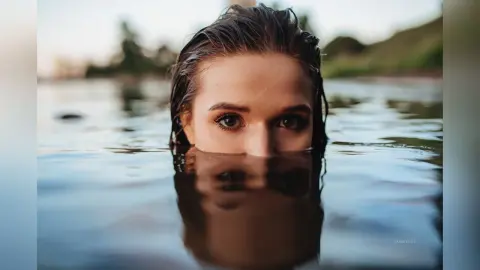 Close-up portrait of a young woman with wet hair peeking out of calm lake water during a serene summer day.