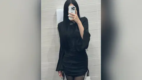 AlettaFlorence poses in a mirror wearing a long-sleeved black ruched dress and long black hair, standing in a tiled bathroom.