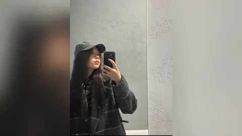 Young woman in a grey cap and dark plaid coat taking a moody mirror selfie in a minimalist room.