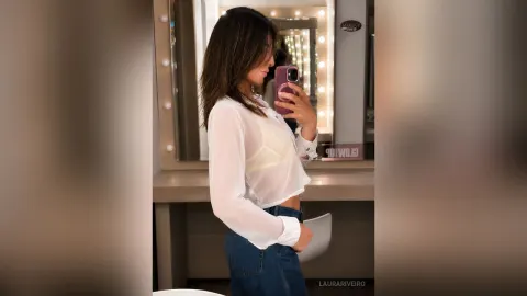 LauraRiveiro — LauraRiveiro takes a mirror selfie in a white sheer shirt and blue jeans while standing in front of a lighted vanity mirror.