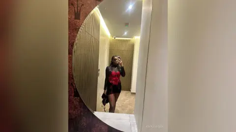 AmyJacobs poses in a mirror wearing a red and black corset top and black shorts, showing off her long dark hair and legs.