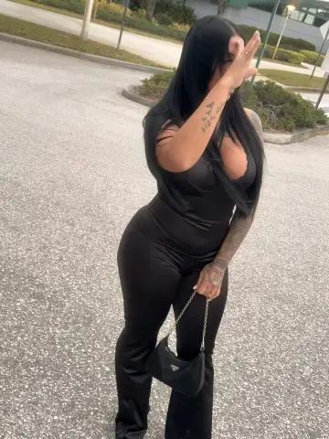 Woman in a sleek black jumpsuit and chain handbag standing on an outdoor paved walkway.