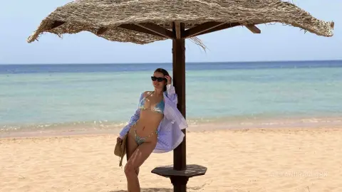 Woman in bikini and blue striped shirt relaxing under a straw umbrella on a sunny tropical beach.