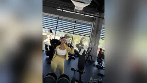 NicoleBella stands in a gym wearing a white sports bra and yellow leggings with a matching yellow jacket and headphones.