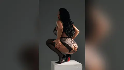 CassyeCross squats on a white block wearing a black leopard print bra and matching thong with black stockings.
