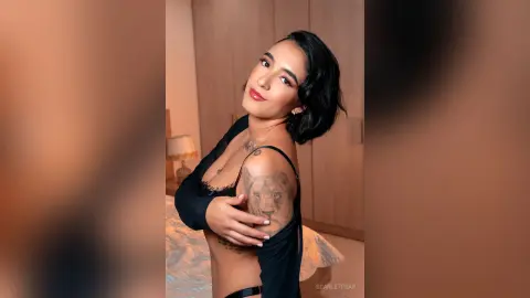 ScarletPear poses with her hand on her chest, showing off her shoulder tattoo and black lace top while looking at the camera.