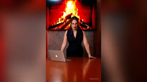 Professional woman in a black sleeveless dress working on a laptop by a cozy glowing fireplace.
