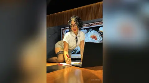 Young woman in a white shirt working on a laptop at a wooden desk in a moody, dimly lit room.