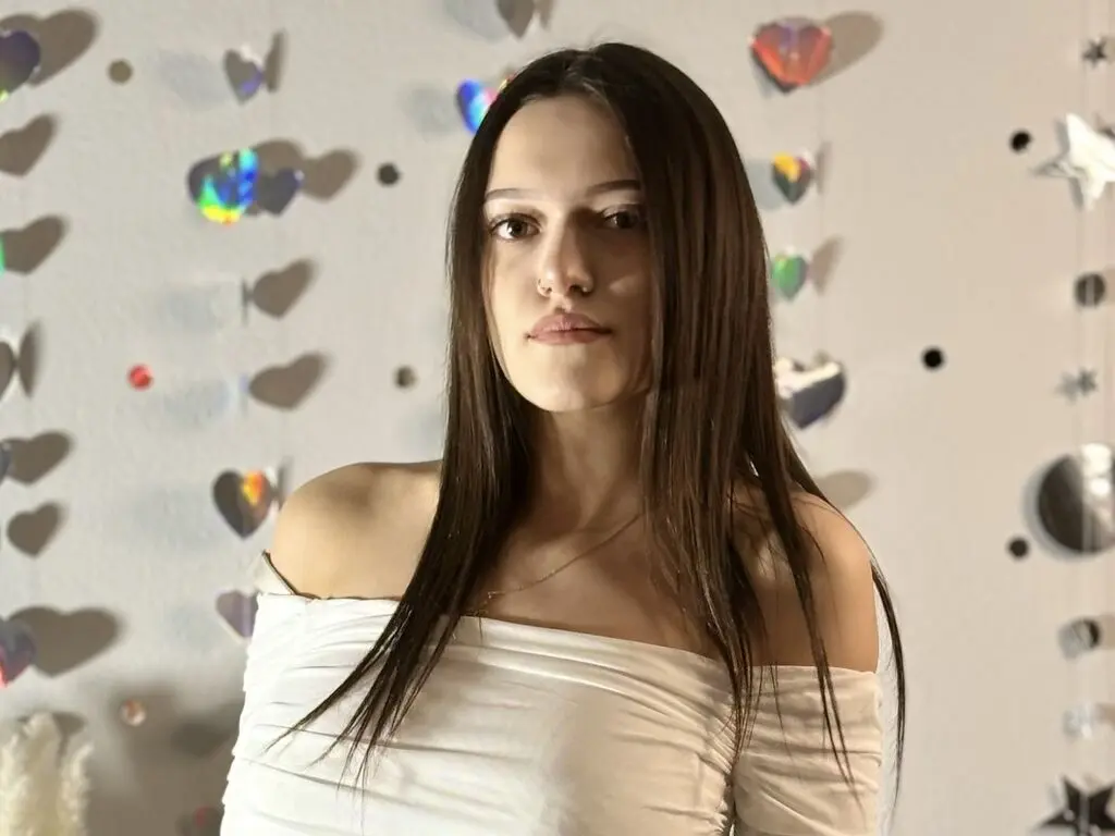 Young woman in a white off-the-shoulder top posing against a wall decorated with heart-shaped disco decorations.