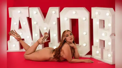 AmberNash — Nude woman lying on red floor in front of large white AMOR marquee lights for a romantic mood.