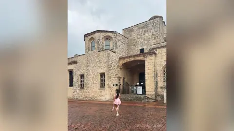 Young woman in a pink dress walking through a historic stone courtyard under a cloudy sky.