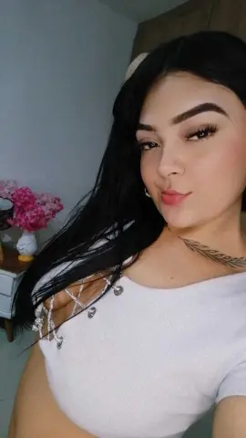 Young woman with long dark hair wearing a white lace crop top in a bright indoor setting.