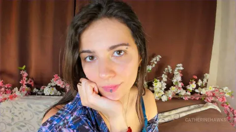 Young woman in a blue patterned top resting her chin on her hand in a cozy room with floral decor.