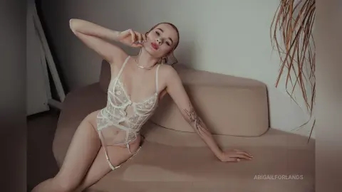 Blonde woman in white lace lingerie relaxing on a beige sofa in a minimalist, bright indoor setting.