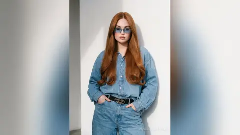 Redheaded woman in denim shirt and sunglasses posing against a white wall in bright sunlight.