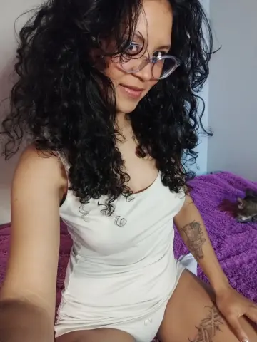 Hathorlove smiles wearing clear glasses and a white tank top, showing off her long, dark curly hair and arm tattoos while sitting on a purple towel.