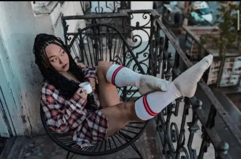 Woman in plaid shirt and white socks relaxing on a balcony chair with a coffee mug on a city morning.