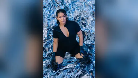 KimRoberth poses in a black bodysuit and black gloves against a shiny silver foil background.
