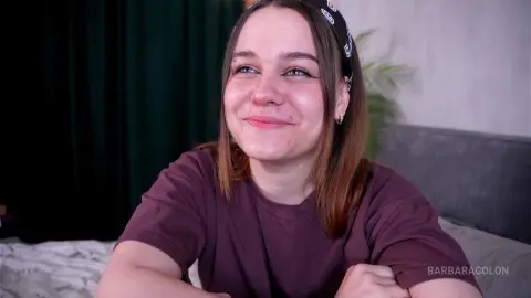 Smiling young woman in a casual purple t-shirt sitting in a cozy, dimly lit bedroom setting.