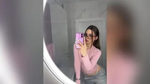 Young woman in a pink long-sleeve top taking a stylish mirror selfie in a modern marble bathroom.