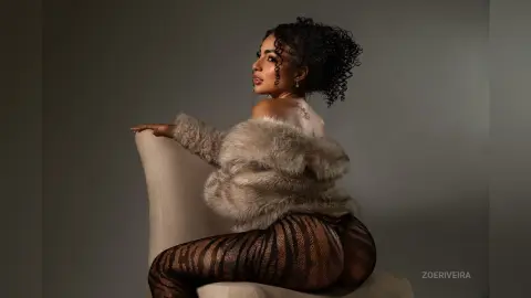 Glamorous woman in a faux fur coat and patterned tights sitting on a chair in a moody studio setting.