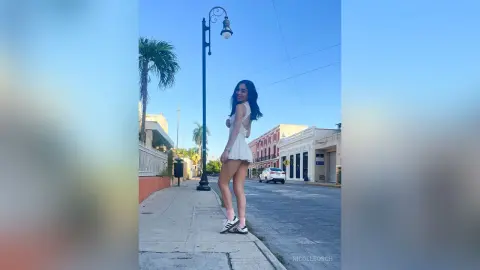 Young woman in a white summer dress walking on a sunny city sidewalk near palm trees.