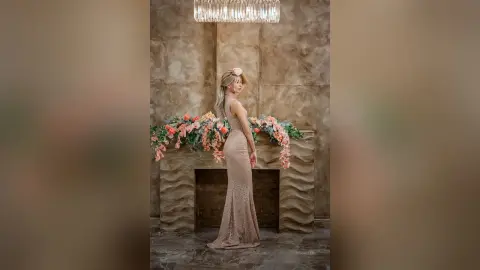 Elegant woman in a shimmering rose gold evening gown posing by a floral mantelpiece in a moody studio setting.