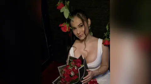 NahomiRomero poses in a white tank top while holding a large bouquet of red roses against a dark background.