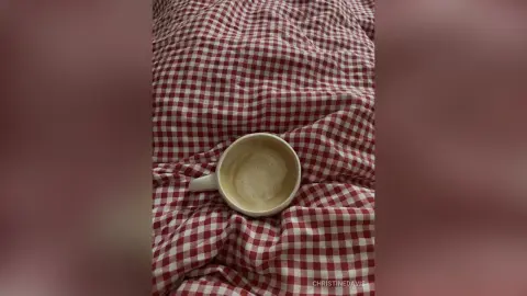 ChristineDavis poses a white mug filled with coffee or tea resting on a red and white checkered cloth or bedsheet.