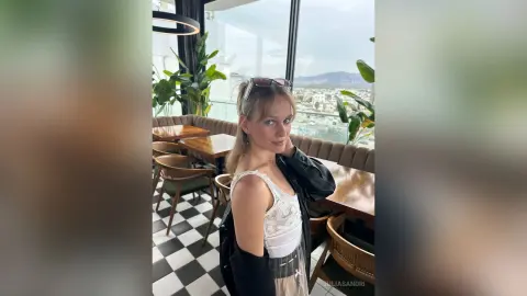 Young woman in a white lace top and black jacket posing in a modern rooftop cafe with a city view.