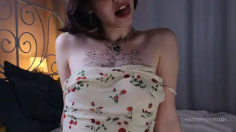 Young woman with chest tattoos in a floral sundress sitting in a dimly lit bedroom.