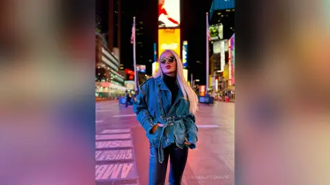 Stylish blonde woman in a denim jacket and sunglasses standing in Times Square at night.