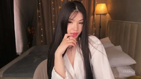Young woman with long dark hair in a white shirt sitting on a bed in a cozy, dimly lit room.